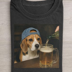 Op de afbeelding: Donkergrijs T-shirt met een beagle met een blauwe denim baseballcap, die kijkt naar een bierpul die wordt gevuld. De hond heeft bruine en witte aftekeningen. Het bier heeft een licht amberkleur.