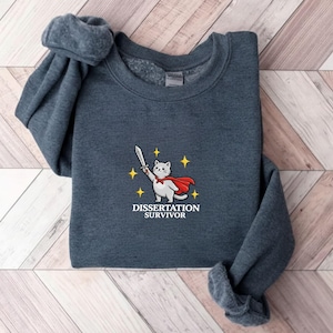 May include: A dark gray sweatshirt featuring an embroidered design of a white cat wearing a red cape and holding a sword. The text "DISSERTATION SURVIVOR" is below the cat. The sweatshirt is folded on a wooden surface.