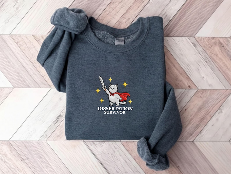 May include: A dark gray sweatshirt featuring an embroidered design of a white cat wearing a red cape and holding a sword. The text "DISSERTATION SURVIVOR" is below the cat. The sweatshirt is folded on a wooden surface.
