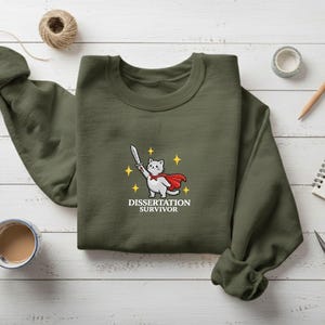 May include: An olive green sweatshirt with a crew neck. The front features a cartoon cat wearing a red cape and holding a sword, with the text "Dissertation Survivor" below. The sweatshirt is surrounded by stationery items.