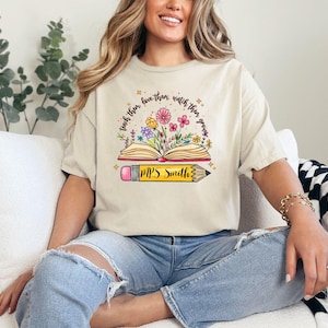 May include: A beige t-shirt with a graphic of an open book with flowers growing out of it. The text "teach them love, then watch them grow" is written around the book. A pencil with the text "Mrs. Smith" is below the book.