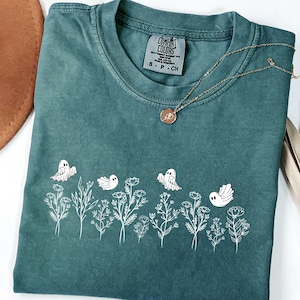 Puede incluir: Camiseta Comfort Colors verde azulado con un diseño floral y fantasma blanco. Un collar de oro con un colgante descansa sobre la camisa. La camisa tiene cuello redondo y mangas cortas. La etiqueta dice "S P CH".