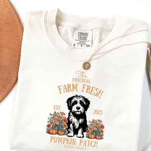 May include: Cream-colored t-shirt with a graphic featuring a black and white dog, pumpkins, and flowers. The text reads "The Original Farm Fresh Pumpkin Patch Open Daily, Est. 2025". A gold necklace and a hat are in the background.