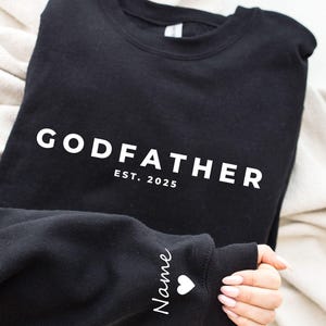 Pu&ograve; includere: Felpa nera con la scritta "GODFATHER" in lettere bianche, con "EST. 2025" sotto. La manica ha il nome "Name" in corsivo con un cuore bianco. La felpa &egrave; piegata su una coperta color crema.