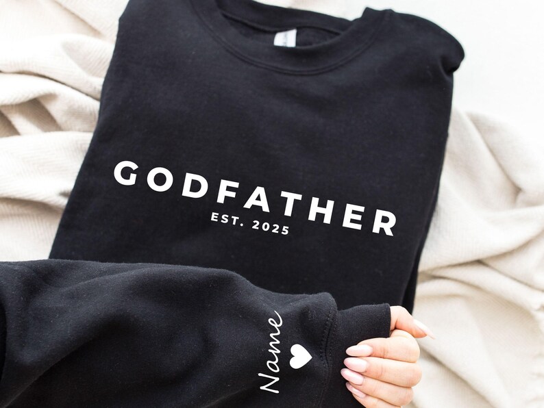 Pu&ograve; includere: Felpa nera con la scritta "GODFATHER" in lettere bianche, con "EST. 2025" sotto. La manica ha il nome "Name" in corsivo con un cuore bianco. La felpa &egrave; piegata su una coperta color crema.