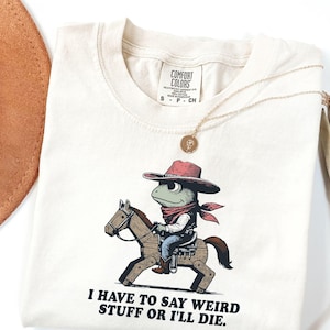 Puede incluir: Camiseta color crema con un dibujo de una rana vaquera montada en un caballo de madera. La rana lleva un sombrero y una bandana rojos. El texto "I HAVE TO SAY WEIRD STUFF OR I'LL DIE" está impreso debajo. También se ven un collar dorado y un sombrero marrón.