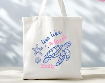 May include: White canvas tote bag with a blue and pink design. The design features a sea turtle, a starfish, and a seashell. The text "live like a local" is written in blue and pink. The name "Emily" is written in pink at the bottom of the bag.