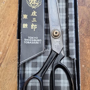 May include: A pair of steel tailor shears with black handles and a brass pivot screw, resting in a black and grey plaid box. The box features gold text reading Tokyo Shozaburo Tobasami and a gold emblem. The metal blades are silver and pointed.