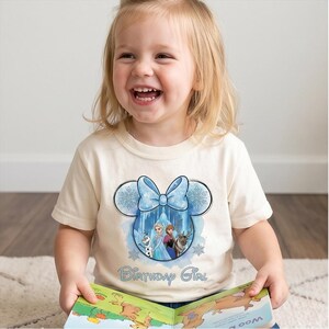 May include: A cream-colored short-sleeve t-shirt featuring a graphic of a blue bow and Mickey Mouse ears silhouette containing characters from Frozen. The design includes snowflakes and the text Birthday Girl in blue script lettering.