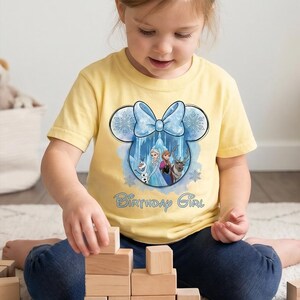May include: A pale yellow short-sleeved t-shirt featuring a graphic of a Mickey Mouse-shaped outline with a light blue bow. Inside the outline are characters from Frozen, including Olaf, Elsa, Anna, and Sven. The text Birthday Girl is printed below.