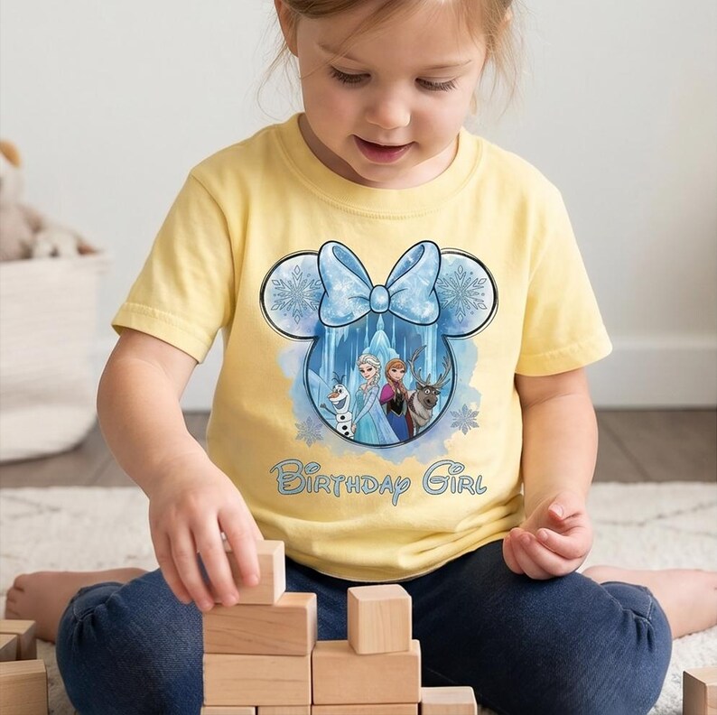May include: A pale yellow short-sleeved t-shirt featuring a graphic of a Mickey Mouse-shaped outline with a light blue bow. Inside the outline are characters from Frozen, including Olaf, Elsa, Anna, and Sven. The text Birthday Girl is printed below.