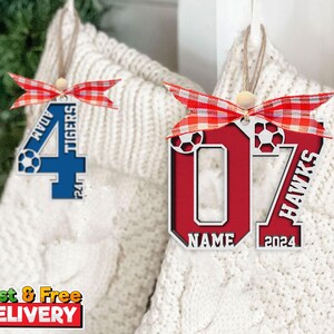 May include: Wooden soccer-themed ornaments with red and blue accents. One features the number 4 with "ADAM" and "TIGERS" text, the other displays the number 07 with "HAWKS" and "NAME 2024" text. Each ornament has a red and white ribbon.
