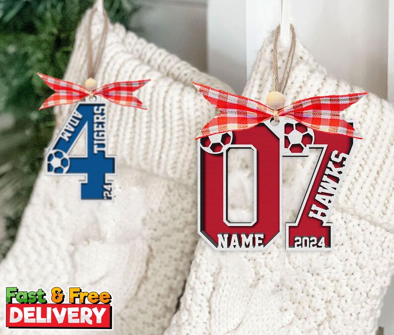 May include: Wooden soccer-themed ornaments with red and blue accents. One features the number 4 with "ADAM" and "TIGERS" text, the other displays the number 07 with "HAWKS" and "NAME 2024" text. Each ornament has a red and white ribbon.