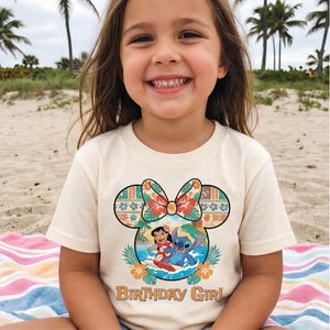 May include: A cream-colored short-sleeve t-shirt featuring a graphic of Lilo and Stitch surfing inside a Mickey Mouse ear silhouette with a floral bow. The design includes tropical leaves, hibiscus flowers, and the text "BIRTHDAY GIRL" in brown.