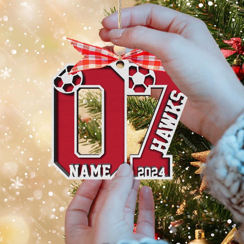 May include: A personalized wooden Christmas ornament in the shape of a jersey. The ornament is red with white accents, featuring soccer ball cutouts, the number 07, and the word "HAWKS". It includes space for a name and the year 2024, and is topped with a red and white checkered bow.