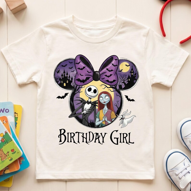 May include: A cream-colored short-sleeve t-shirt featuring a graphic of Jack Skellington, Sally, and Zero inside a Mickey Mouse-shaped silhouette. The design includes a purple bow, bats, a moon, and the text "BIRTHDAY GIRL" in black lettering.