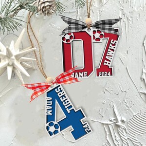 May include: Two personalized soccer-themed ornaments. One is red with the number 07, the name "NAME", and "HAWKS 2024". The other is blue with the letter "A", the name "ADAM", and "TIGERS 24". Each has a soccer ball design.