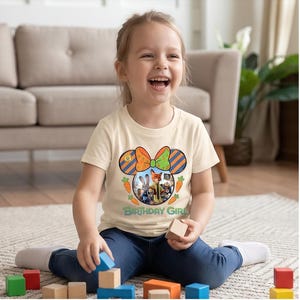 K&ouml;nnte beinhalten: Ein cremefarbenes Kurzarm-T-Shirt mit einer Grafik einer Mausohren-Silhouette mit orangefarbenen und blauen Streifen, einer gr&uuml;nen und orangefarbenen Schleife sowie Zootopia-Figuren. Der Text "BIRTHDAY GIRL" ist darunter in Gr&uuml;n gedruckt.