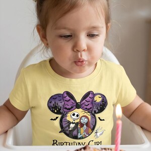 May include: A pale yellow short-sleeved t-shirt featuring a graphic of Jack Skellington and Sally from The Nightmare Before Christmas inside a Mickey Mouse ear silhouette with a purple bow, bats, a moon, and the text Birthday Girl.