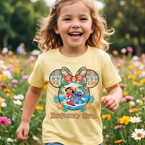 May include: A light yellow t-shirt featuring a graphic of Lilo and Stitch surfing inside a Mickey Mouse ear silhouette. The design includes a floral bow, tropical leaves, and the text "BIRTHDAY GIRL" printed in a brown retro-style font.