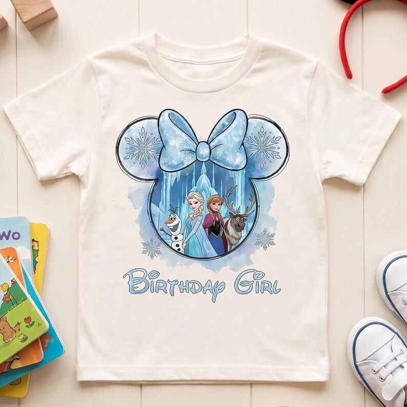 May include: A cream-colored short-sleeve t-shirt featuring a graphic of Mickey Mouse ears filled with an icy blue scene of characters from Frozen, including Olaf, Elsa, Anna, and Sven, topped with a blue bow and the text Birthday Girl.