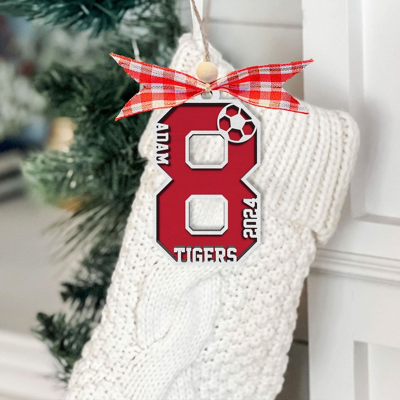 May include: A red and white soccer-themed Christmas ornament shaped like the number 8. The ornament has the name "ADAM", the year "2024", and the word "TIGERS" printed on it. It is decorated with a red and white plaid ribbon.