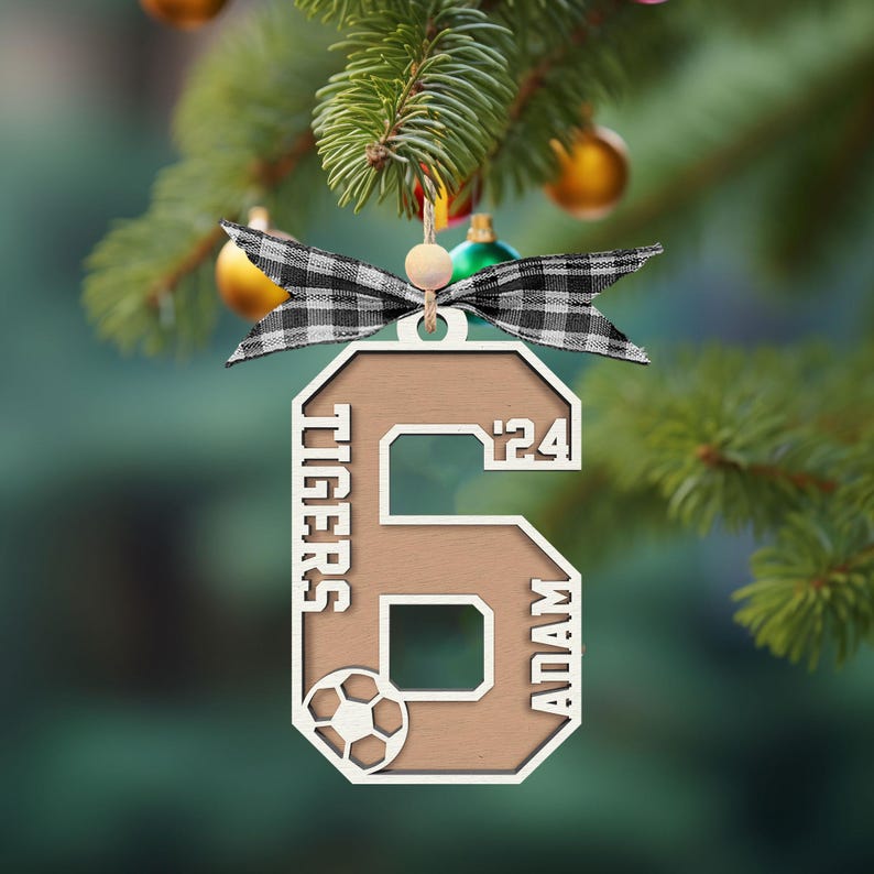 May include: A wooden Christmas ornament in the shape of the number 6, with the words "TIGERS" and "ADAM" and the year "'24" inscribed. A soccer ball graphic is included. The ornament is decorated with a black and white plaid bow.