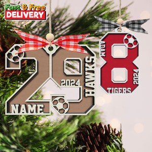 May include: Two wooden soccer ornaments hanging from twine. One is brown with the number 28, the word "NAME", and "2024". The other is red with the number 8, the word "TIGERS", and "2024". Both have soccer ball cutouts.
