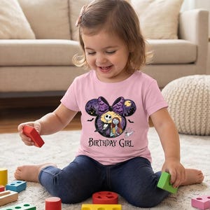 May include: A light pink short-sleeved t-shirt featuring a graphic of Jack Skellington and Sally inside a Mickey Mouse-shaped silhouette with a purple bow, bats, and a moon. The text Birthday Girl is printed in black below the graphic.