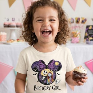 May include: A cream-colored short-sleeve t-shirt featuring a graphic of Jack Skellington and Sally from The Nightmare Before Christmas inside a Minnie Mouse ear silhouette with a purple bow, bats, and the text Birthday Girl printed below.