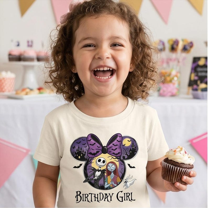 May include: A cream-colored short-sleeve t-shirt featuring a graphic of Jack Skellington and Sally from The Nightmare Before Christmas inside a Minnie Mouse ear silhouette with a purple bow, bats, and the text Birthday Girl printed below.
