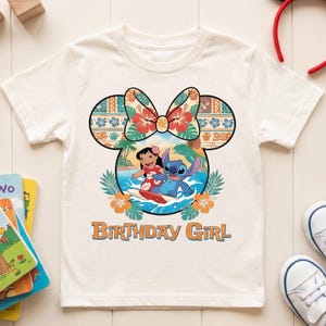 May include: A cream-colored short-sleeve t-shirt featuring a graphic of Lilo and Stitch surfing inside a Mickey Mouse ear silhouette with a floral bow. The design includes orange hibiscus flowers and the text Birthday Girl in orange letters.