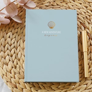 Könnte beinhalten: Ein hellblaues Notizbuch mit einem goldenen Muschel-Design und dem Text "A new adventure begins".