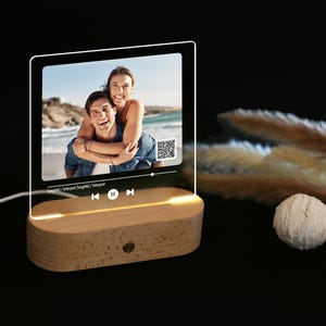 Könnte beinhalten: Eine transparente Acryl-Fotodisplay mit einem Foto eines Paares am Strand, auf einem Holzsockel. Das Display hat eine Musikplayer-Oberfläche und einen QR-Code. Der Sockel wird von einem warmen Licht beleuchtet.