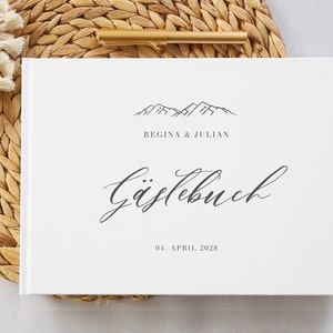 Könnte beinhalten: Weißes Gästebuch mit einer Bergkette-Illustration und dem Text "Regina & Julian" und "Gästebuch" in Schreibschrift. Das Datum "04. APRIL 2028" ist unter dem Text in Schreibschrift gedruckt.