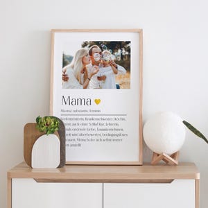 Puede incluir: Una impresión enmarcada con una foto de una familia soplando burbujas. La impresión tiene la palabra "Mama" en letras grandes con un corazón y una definición de la palabra en texto más pequeño.