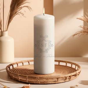 Könnte beinhalten: Weiße Säulenkerze mit einem grauen floralen Design und den Initialen "SF" in einem Schild. Der Text "Sarah und Fabian" ist unter dem Design gedruckt.