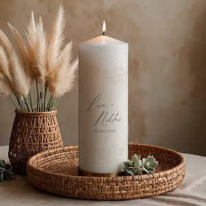 Könnte beinhalten: Weiße Kerze in Säulenform mit einem floralen Muster und dem Text "Lisa & Niklas 18. JULI 2027" an der Seite. Die Kerze steht auf einem geflochtenen Tablett.