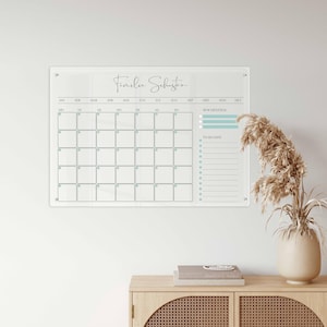 Könnte beinhalten: Ein klarer monatlicher Acrylkalender mit dem Text "Familu Schuster" in Schreibschrift. Das Kalenderraster ist hellblau, mit einer To-Do-Liste und einem Wochenziele-Bereich. Der Kalender ist an der Wand befestigt.