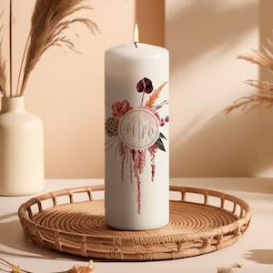 Könnte beinhalten: Weiße Säulenkerze mit einem floralen Design und den Initialen "HM" in einem Kreis. Das Blumenmuster enthält rosa, orange und burgunderfarbene Blumen.