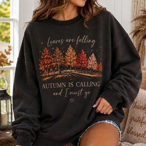 May include: A charcoal gray crewneck sweatshirt featuring a graphic of a row of autumn trees in shades of orange, red, and yellow. The text reads, "Leaves are falling, AUTUMN IS CALLING and I must go" with a small heart icon below the lettering.