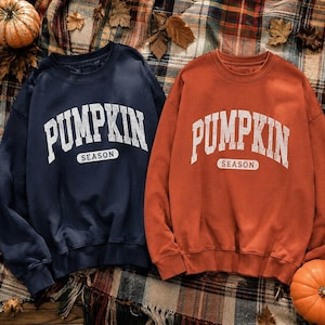 May include: Two crewneck sweatshirts, one in navy blue and one in burnt orange, each featuring the white distressed text PUMPKIN SEASON printed in an arched collegiate font across the chest with long sleeves and ribbed cuffs and hem.