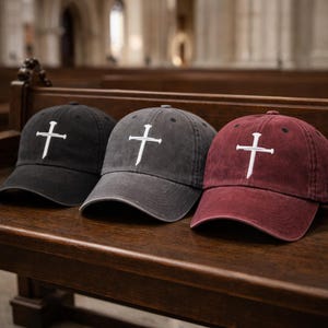 May include: Three baseball caps in black, gray, and burgundy, each embroidered with a white cross. The caps are displayed on a wooden bench, with a church interior in the background.