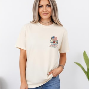 May include: Cream-colored t-shirt featuring a small graphic on the upper left chest. The graphic includes an American flag, flowers, and text that reads "250th Anniversary". The shirt is paired with blue jeans.