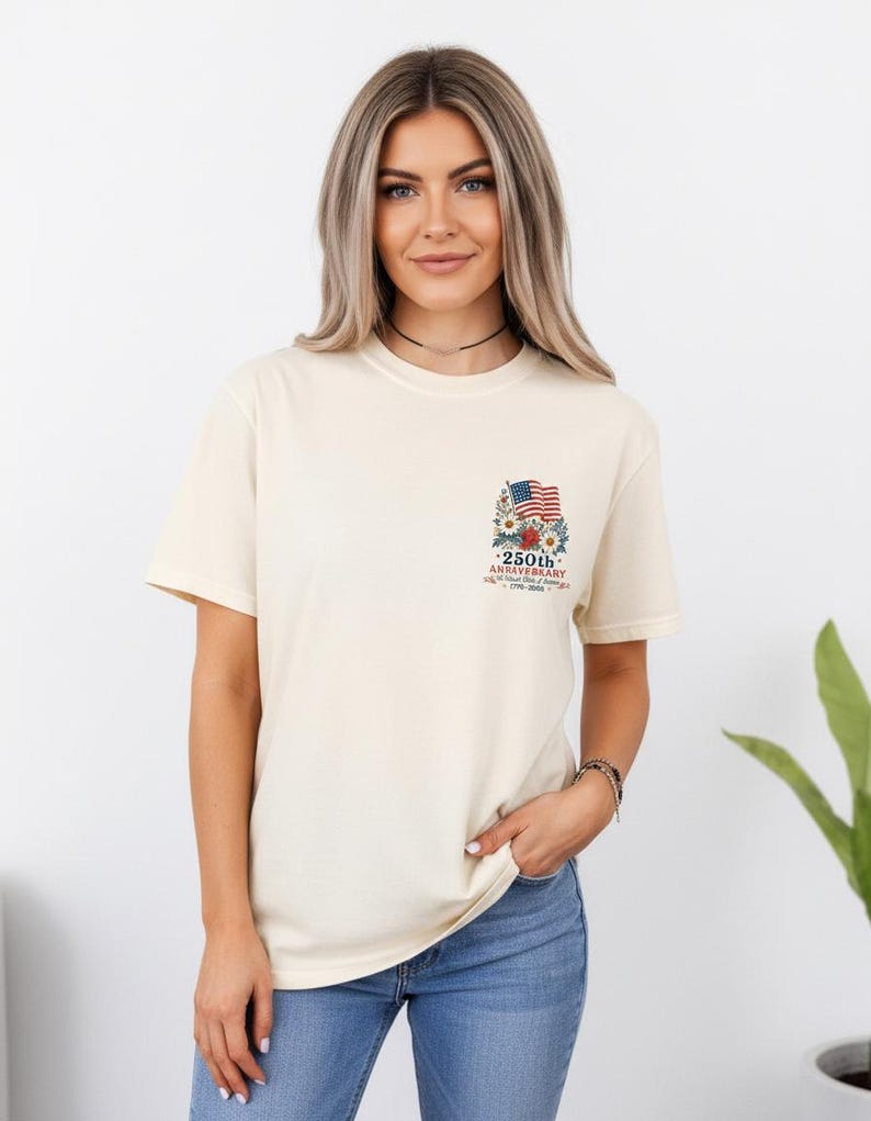May include: Cream-colored t-shirt featuring a small graphic on the upper left chest. The graphic includes an American flag, flowers, and text that reads "250th Anniversary". The shirt is paired with blue jeans.