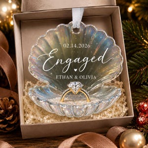 May include: An iridescent, shell-shaped ornament featuring the text 02.14.2026, Engaged, and ETHAN & OLIVIA. A gold-toned ring with a large clear stone sits in the center of the shell, which is filled with small white granules and hangs by a white ribbon.