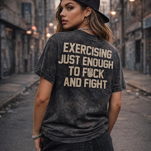 May include: A dark gray t-shirt with the text "EXERCISING JUST ENOUGH TO FUCK AND FIGHT" in a bold, off-white font. The shirt is worn with a black baseball cap and dark jeans. The image is taken outdoors.
