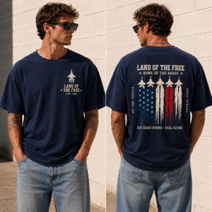 May include: Navy blue t-shirt with a graphic of a jet and the text "LAND OF THE FREE 1776-2026" on the front. The back features an American flag design with jets and the text "LAND OF THE FREE HOME OF THE BRAVE 250 YEARS STRONG STILL FLYING".