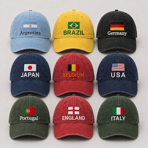 May include: Nine baseball caps in various colors, each embroidered with a country's flag and name. The caps include Argentina, Brazil, Germany, Japan, Belgium, USA, Portugal, England, and Italy. The caps are made of a soft, worn fabric.