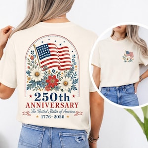 May include: Cream-colored t-shirt featuring a patriotic design. The back displays an American flag with floral accents, the text "250th Anniversary The United States of America 1776-2026". A smaller flag design is on the front.